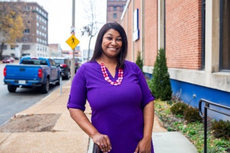 Woman in purple dress smiling while leaning on a railing on sidewalk. 